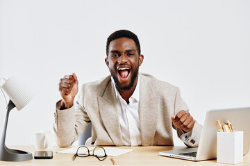 Happy young African American man in beige blazer celebrates success at office desk, smiling and...