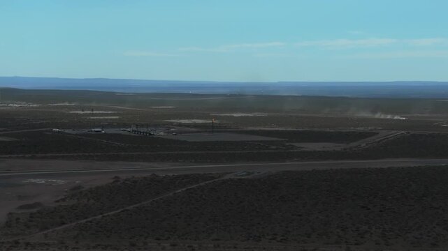 Vaca Muerta plateau. A car appears in the distance, kicking up dust from the road. A fracking drill is visible in the distance. 4K drone footage.