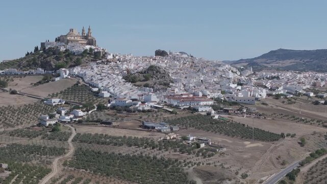 Wide aerial drone view of Olvera white hill town in Andalusia, Spain, featuring historic castle and church above a dense whitewashed village, with olive groves, farmland and Mediterranean countryside