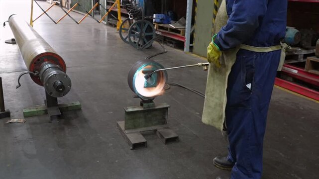 Metal fabrication workshop showing worker in coveralls, gloves and apron using gas torch to heat one end of rotating cylindrical metal pipe supported on stands with glowing metal indicating preheat