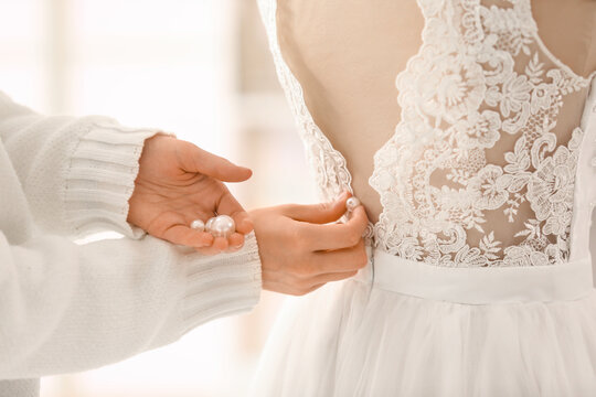 Bride getting ready with helper fastening lace wedding dress buttons, close up of elegant bridal gown back detail and pearls in hand, romantic preparation before ceremony