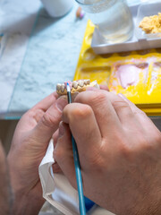 Ceramic tooth crown being painted by dental technician with pink gum layer on dental model. Manual restoration process and craftsmanship in prosthetics lab.