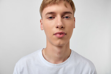 Close up of a young man with light hair wearing a white t shirt against a plain background,...