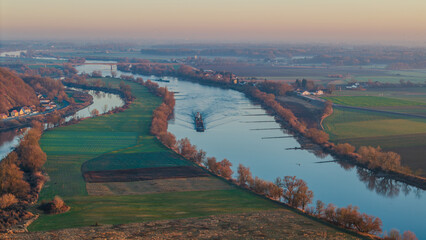Schiff f&auml;hrt den Fluss bei Sonnenuntergang entlang, umgeben von B&auml;umen und Wiesen