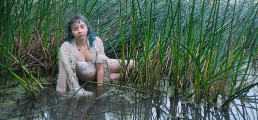 Young woman sitting in shallow lake among tall reeds wearing light lace dress gazing at camera with calm presence expressing nature bond vulnerability quiet strength and contemplative mood