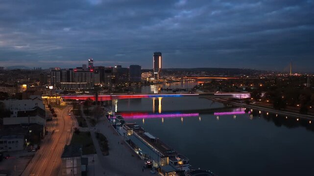 Aerial drone view of Belgrade Waterfront at blue hour, showcasing the illuminated Sava River, modern skyline, Ada Bridge, and colorful reflections on the water
