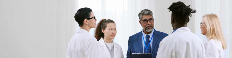 Group of diverse young adult and middle aged doctors and medical professionals standing together, discussing patient case with senior Caucasian man holding clipboard leading conversation