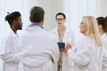 Fototapeta premium Group of diverse middle aged doctors standing in discussion, Caucasian woman with short dark hair holding clipboard leading conversation, medical professionals collaborating