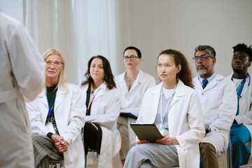 Group of diverse middle aged and young adult men and women doctors listening attentively to speaker during medical seminar, multiethnic professionals wearing lab coats seated indoors