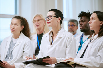 Fototapeta premium Group of multiethnic young adult and middle aged men and women doctors listening attentively during medical seminar, holding notebooks and pens, sitting together in clinical setting