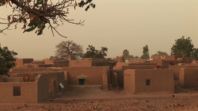 Traditional mud-brick compound houses in a dusty Sahel village near Navrongo, northern Ghana, with flat roofs, tiny windows, scattered trees and a hazy late-afternoon sky.