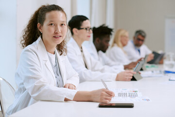 Portrait of young adult Asian woman sitting at conference table writing on document, multiethnic group of middle aged men and women in lab coats working with digital tablets nearby