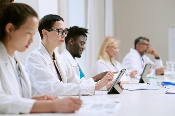 Fototapeta premium Group of multiethnic middle aged and young adult men and women sitting at table using digital tablets and writing, wearing lab coats, participating in professional medical meeting