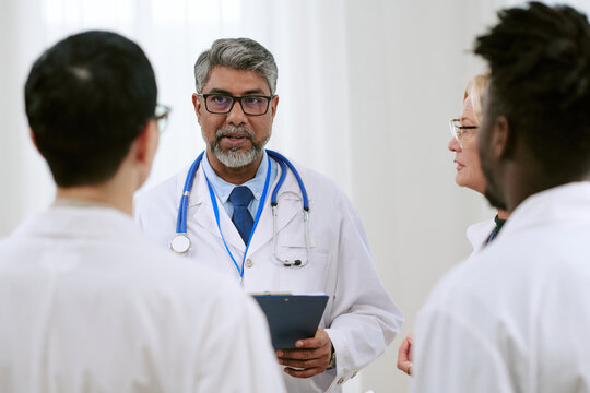 Middle aged South Asian male doctor holding clipboard, addressing diverse group of medical professionals including Caucasian woman and Black man during team discussion in hospital