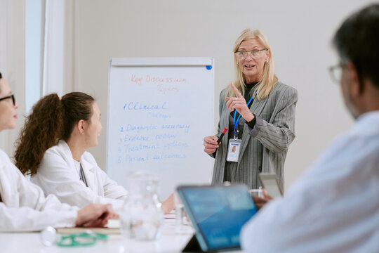 Middle aged Caucasian woman leading medical meeting, gesturing while standing near flip chart with key discussion topics, diverse group of young adult doctors listening and taking notes