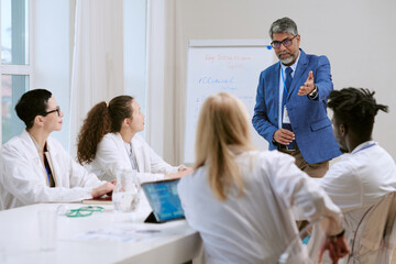 Obraz premium Middle aged Caucasian man standing and gesturing while leading discussion with diverse group of young adult and middle aged doctors sitting at table, listening and taking notes
