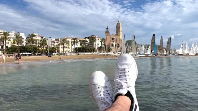 POV Point of view of one person enjoying a summer day with legs outstretched over the water, overlooking the Sitges coastline with its iconic Church of Sant Bartomeu and numerous sailboats in the bay