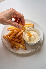 Hand Dipping Golden French Fry into Creamy Mayonnaise on White Plate