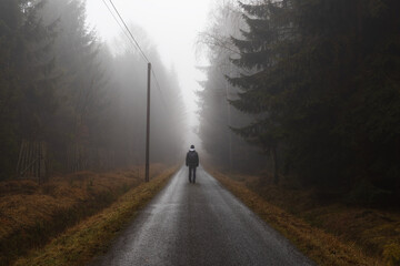 Man with backpack is walking on road through forest at fog morning. Czech landscape