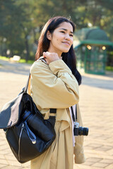 Obraz premium Young photographer in a park wearing a beige trench, bag over shoulder with camera, warm sunlight, exploring outdoor scenes for creative street photography and natural light shots