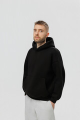 Man stands in a black hoodie with hands in pockets against a plain background during a studio session in daytime