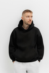 Man stands with hands in pockets wearing a black hoodie in a simple setting with a plain background