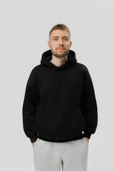 Young man stands with hands in pockets wearing black hoodie and light pants against plain background during indoor session