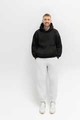 Young man stands in black hoodie and gray sweatpants in a bright indoor space with white background