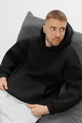Man sits on a couch wearing a black hoodie and light gray pants in a relaxed indoor setting