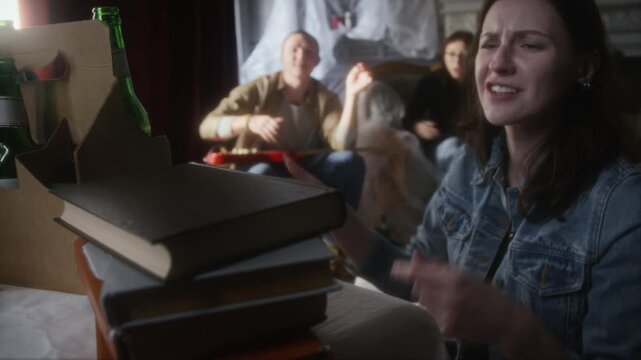 Handheld shot of young woman opening books stacked on table while exploring abandoned house and hanging out with group of young people in hideout, copy space
