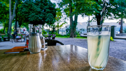 A refreshing drink sits on a wooden table in an outdoor café. Lush green trees surround the area,...