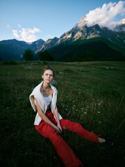 Fototapeta premium Woman sitting in a meadow with red pants and white top, rugged mountains in the background, cloudy sky, outdoor fashion mood, serene landscape and nature inspired scene