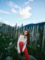 Obraz premium Woman in cozy sweater and red pants sits by a rustic fence in a green hillside landscape, capturing a quiet countryside mood for lifestyle, travel and outdoor photography