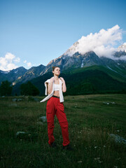 Fototapeta premium Woman in red pants stands in a wide meadow with towering mountains, bright sky, and a light jacket, conveying bold adventure and freedom in nature. Her confident pose suggests exploration, fitness,