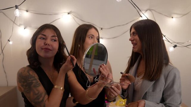 Three happy young female friends putting on makeup together at home while getting ready for a new year's eve party, laughing and talking in a room decorated with festive string lights