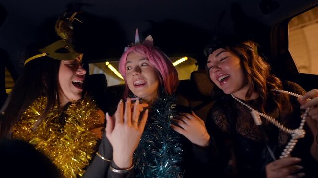 Three beautiful smiling friends in colorful costumes and tinsel garlands are dancing, singing, and having fun together in the backseat of a car during a night city ride to a party