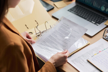 Young adult woman holding document in transparent sheet protector, reviewing paperwork at desk with open binder, laptop and notebook, focusing on business or administrative task