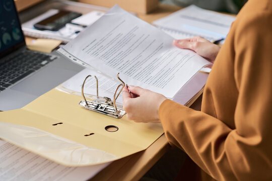 Young adult woman organizing documents in office setting, holding printed papers and placing them into ring binder folder, working at desk with laptop and paperwork visible