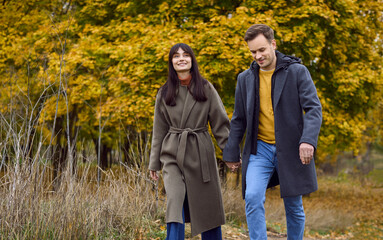 Obraz premium Couple in autumn park walk together smiling. Young adults hold hands beneath golden leaves on a gentle path. Soft natural light and cozy coats add warmth. Concept: love, romance, connection.