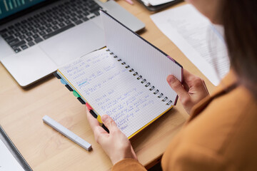 Young adult woman holding open notebook with handwritten notes, sitting at desk with laptop and documents, reviewing information and preparing for work or study