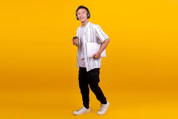 Young Asian Man Holding Disposable Cup And Carrying Laptop While Walking Against Yellow Background