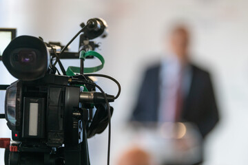 TV camera recording speaker at formal press conference. Highlights media coverage, professional setup, and focused communication setting.