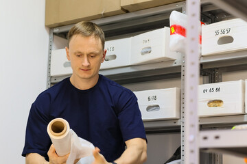 Man in blue shirt holding roll of plastic wrap near shelves with numbered boxes