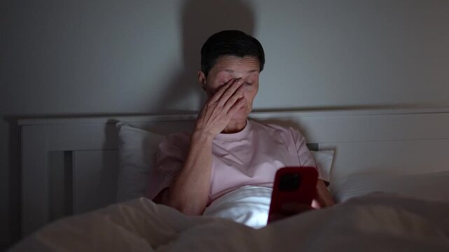 Senior woman with insomnia wasting time in bed on smartphone, lady rubbing eyes and scrolls social media, unhealthy habit using phone before sleep,, bright screen light leading to eyesight issues..