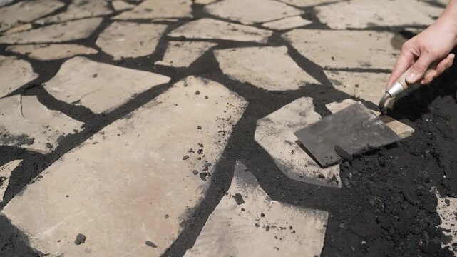 Professional paver using a trowel to apply black grout or sand between irregularly shaped flagstones for a garden path or patio