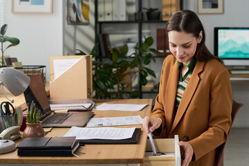 Young adult Caucasian woman sitting at office desk opening drawer and organizing workspace, surrounded by paperwork and digital devices, working in modern workspace