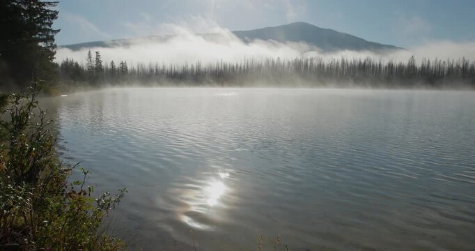 Light morning mist drifts above a quiet lake in a remote setting. Trees edge the shoreline while low clouds settle across the distant landscape, creating a calm, atmospheric scene.