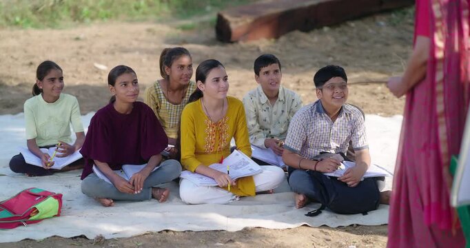 Indian young adult female tutor talk speak share good news to boy girl sitting on mat rural area. Happy child group do study clap hands enjoy have fun joy open air class field day time ground place