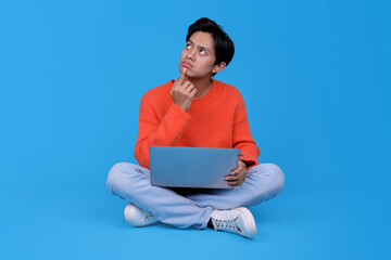 Thoughtful young student with laptop sitting cross-legged on blue studio background