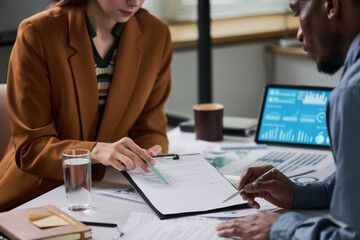 Obraz premium Caucasian young adult woman and Black young adult man reviewing financial documents together at desk, both focusing on paperwork with digital tablet displaying charts in background